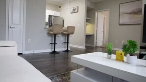 Un salon moderne avec un canapé blanc, une table basse et deux tabourets de bar. On aperçoit à l’arrière-plan un coin cuisine avec des placards et un réfrigérateur.