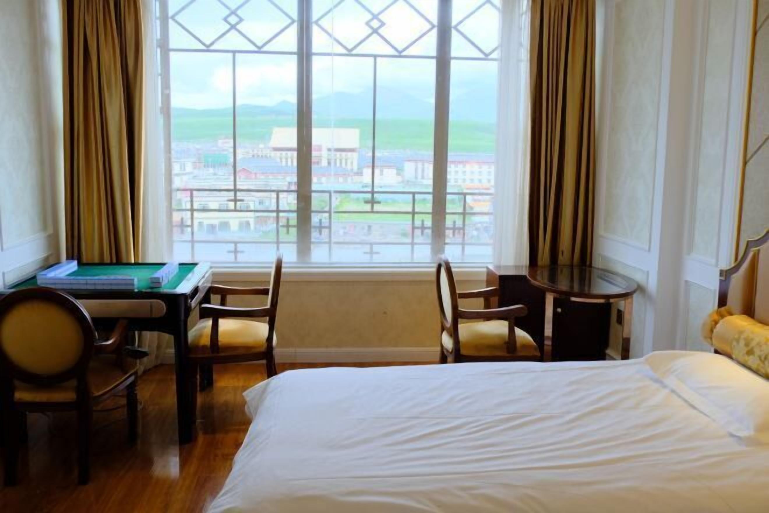 A hotel room with a bed, a desk with a green table, two chairs, and a view of a town through large windows.