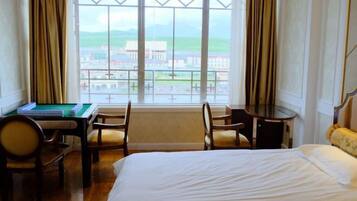 A hotel room with a bed, a desk with a green table, two chairs, and a view of a town through large windows.