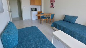 Een kleine, schone kamer met twee eenpersoonsbedden, een kitchenette, een eettafel met stoelen en een deur naar een andere kamer.