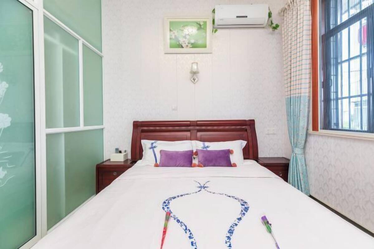 Een keurig opgemaakt bed met decoratieve kussens, een nachtkastje met een lamp, een raam met gordijnen en een airconditioner.