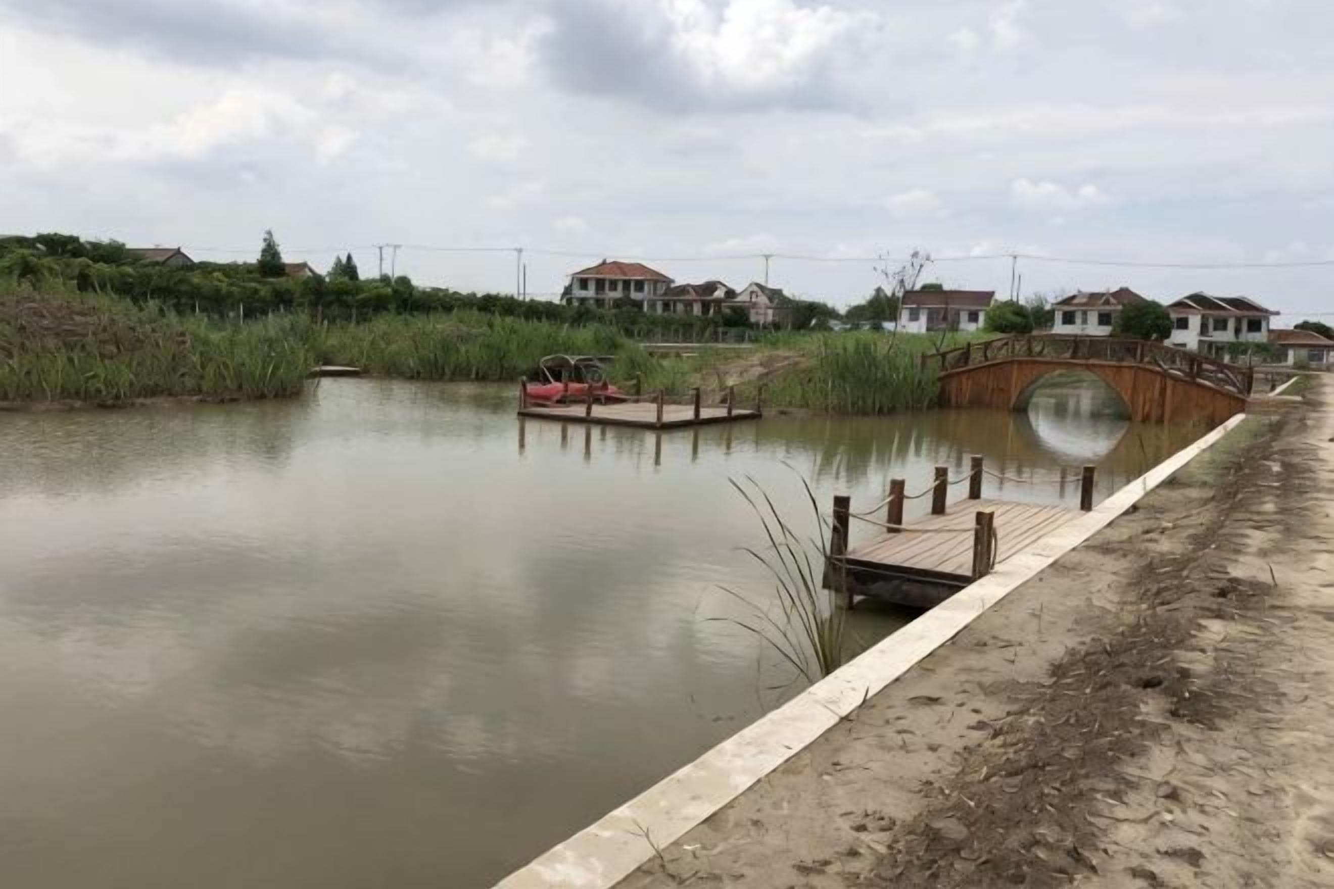 A calm water body with a wooden bridge, a sandy shore, and houses in the background.