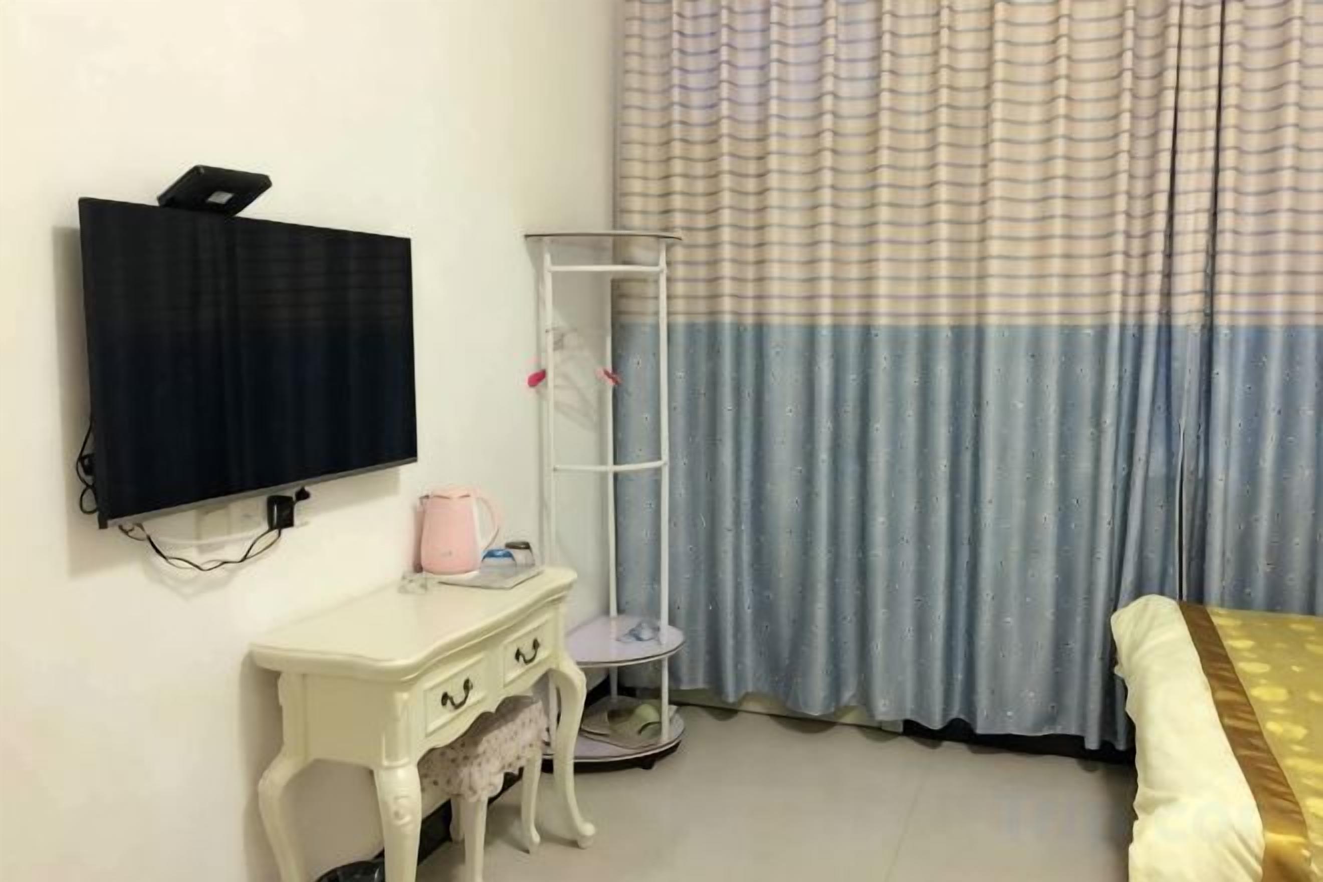 Une chambre dotée d’un téléviseur à écran plat fixé au mur, d’une petite table blanche avec des tiroirs, d’un grand lampadaire et d’un lit recouvert d’une couvre-lit blanche et dorée.