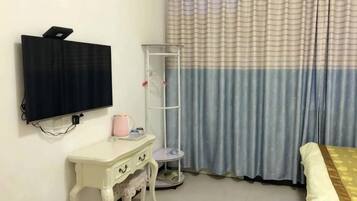 Une chambre dotée d’un téléviseur à écran plat fixé au mur, d’une petite table blanche avec des tiroirs, d’un grand lampadaire et d’un lit recouvert d’une couvre-lit blanche et dorée.