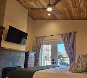 A bedroom with a bed, a flat-screen TV, a ceiling fan, and a stone fireplace. - Lake Coeur d'Alene Inn Guest House (Coeur d'Alene)