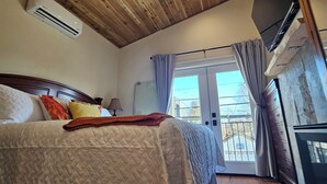 A bedroom with a wooden ceiling, a bed with a quilted bedspread, a ceiling fan, an air conditioning unit, a television, and a view of the outside through a window with curtains. - Lake Coeur d'Alene Inn Guest House (Coeur d'Alene)