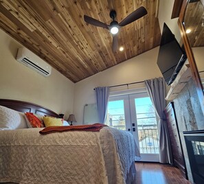 Un dormitorio con techo de madera, cama con colcha acolchada, ventilador de techo, equipo de aire acondicionado, televisor y vistas al exterior a través de una ventana con cortinas.