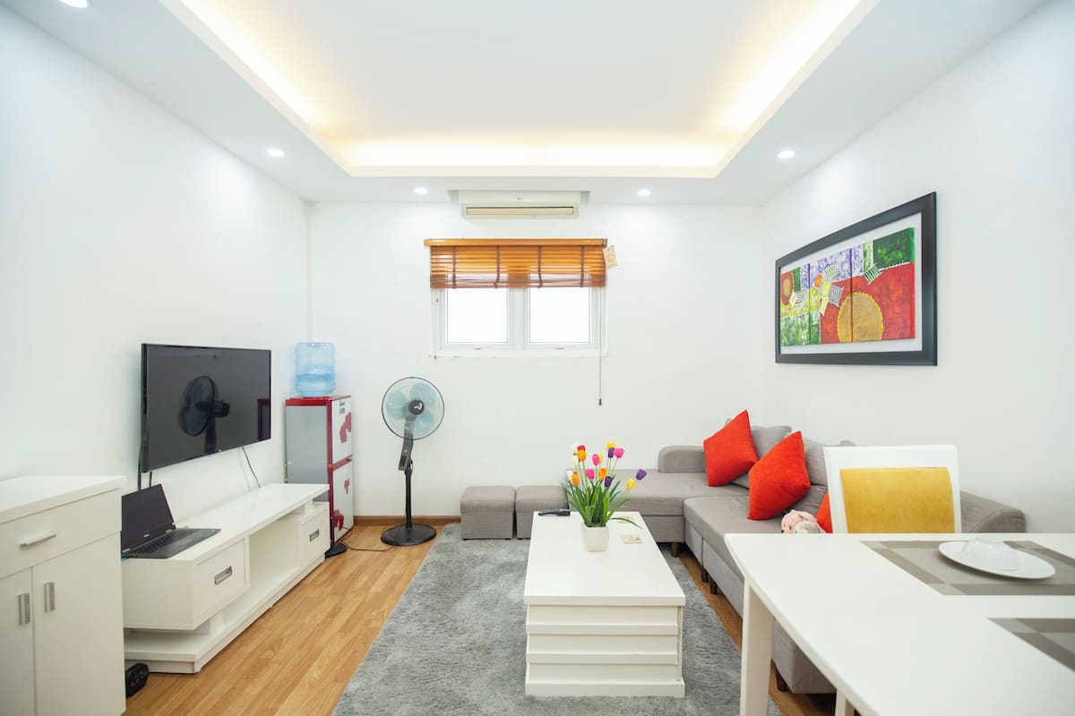 A modern living room with a flat-screen TV, a standing fan, a water dispenser, a sofa with red cushions, a coffee table with a flower vase, and a colorful painting on the wall.