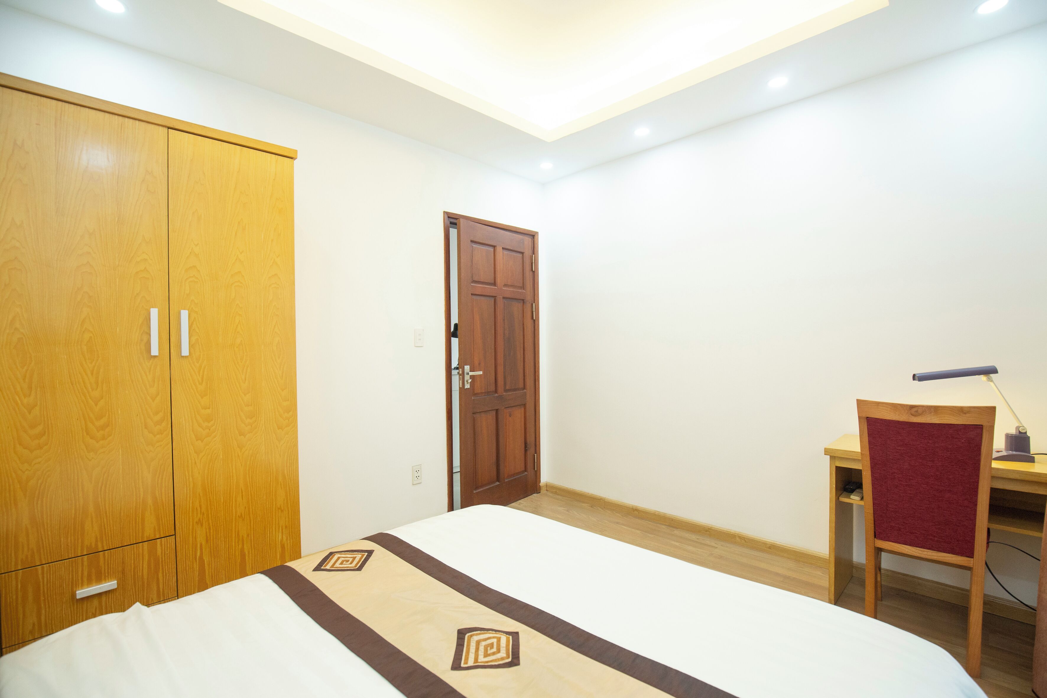 A bedroom with a bed, wooden wardrobe, desk, chair, and a door.