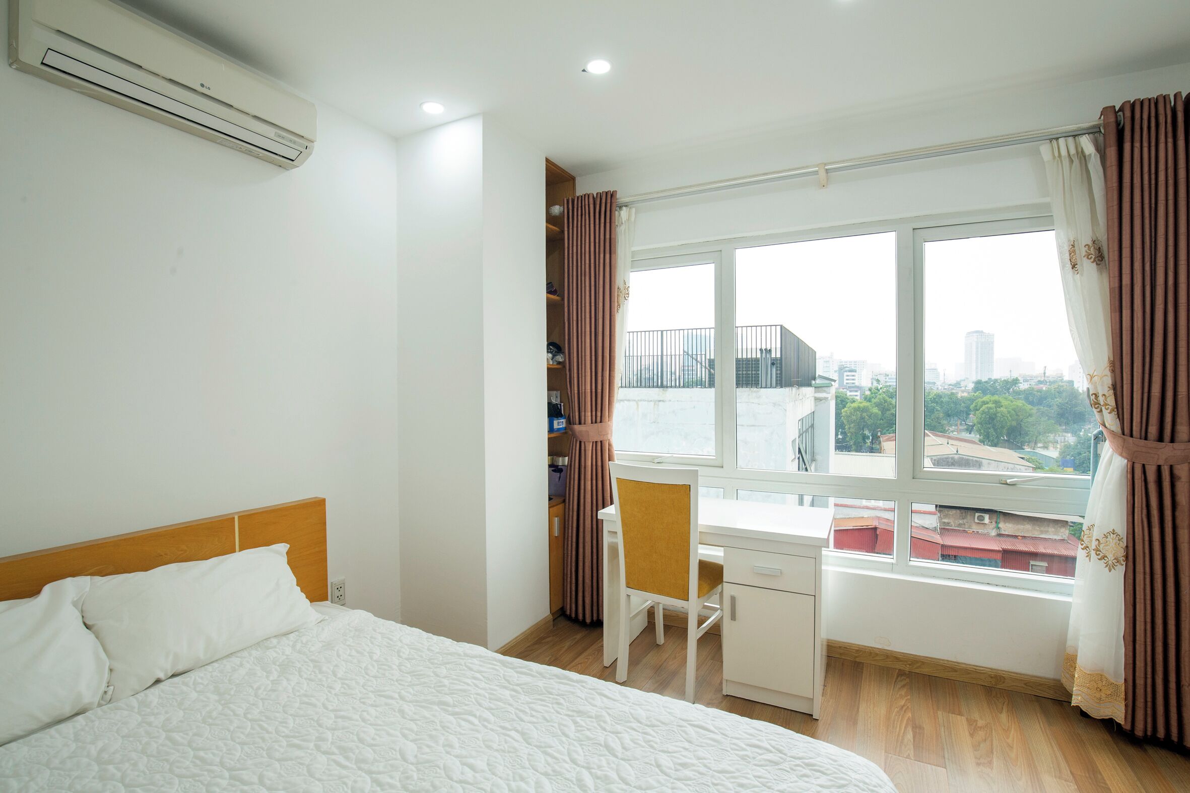 A bedroom with a bed, a desk, and a chair. There is a window with curtains, and an air conditioning unit on the wall.