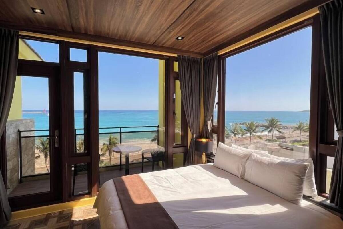 Habitación de hotel con una cama grande, un balcón con vistas a la playa y una mesa con sillas.