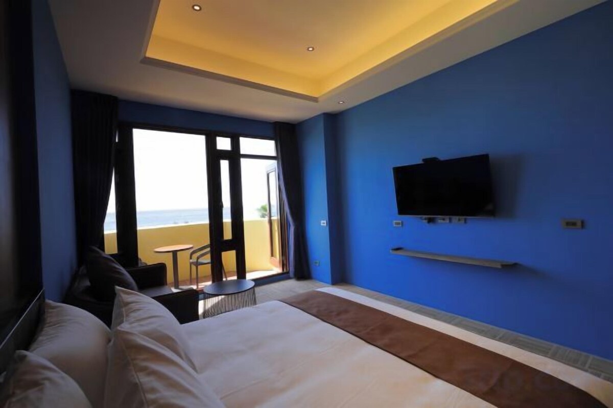 A hotel room with a bed, a TV, a balcony with a table and chairs, and a view of the sea.