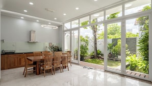 A modern kitchen with wooden cabinets, a dining table, and chairs. It has large sliding glass doors leading to a garden with plants and a tree. - Thao Dien Garden Villa 5BR Luxury Oasis By An Gia Hospitality (Ho Chi Minh City)