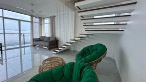Een ruime woonkamer met een hoge plafond, een moderne trap, uitzicht door grote ramen en een groene, beklede fauteuil.