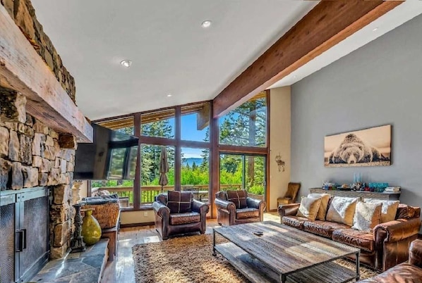 A spacious living room with a stone fireplace, leather furniture, large windows, and a view of greenery.