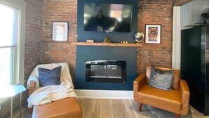 Un salon avec un mur en briques, un grand téléviseur à écran plat fixé au-dessus d’une cheminée, deux canapés en cuir avec des coussins et un plancher en bois.