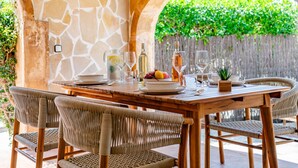 An outdoor dining area with a stone wall, wicker chairs, and a table set for two with wine glasses, a pitcher, and fruit. - Cofitsa Plus by Interhome (Jávea)