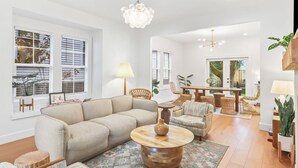 A spacious living room with a beige sofa, round wooden coffee table, and wicker chairs. There is a dining area with a wooden table and chairs, and a large window with blinds. - Old-Portland Charmer in the Alphabet District (Portland)