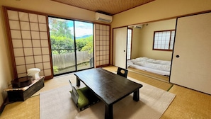 Une pièce de style japonais traditionnel, avec un sol recouvert de tatamis, une table basse, des portes coulissantes et une vue sur la verdure à l’extérieur.