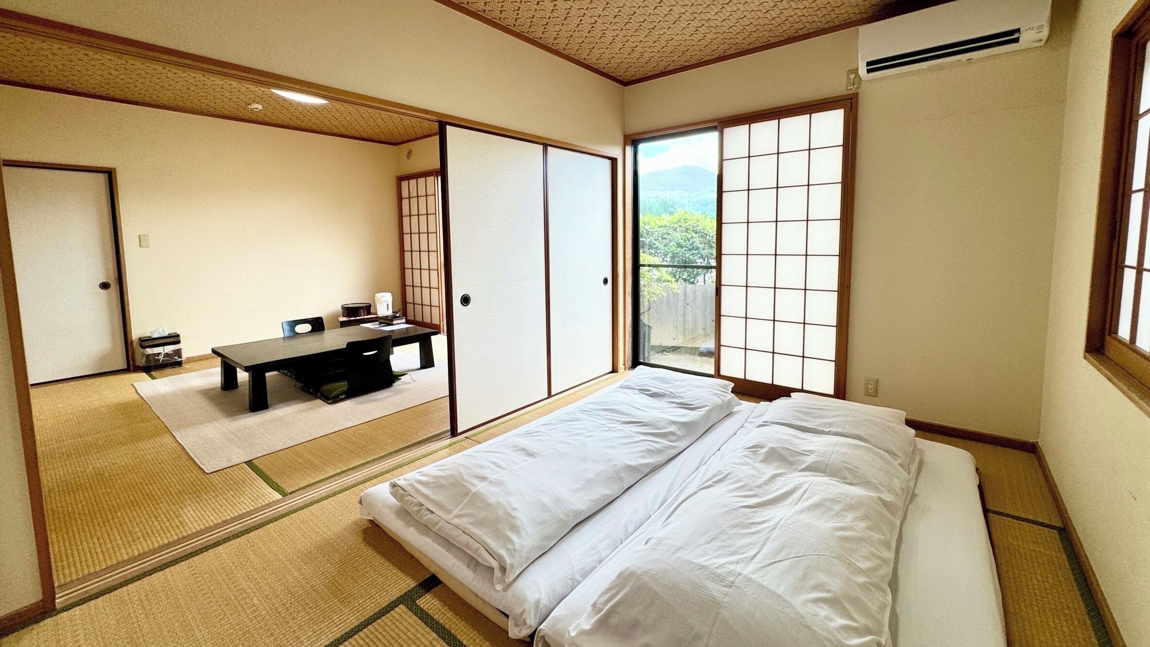 Um quarto japonês tradicional com tatames, futon, mesa baixa com uma cadeira, portas deslizantes e uma grande janela com vista.