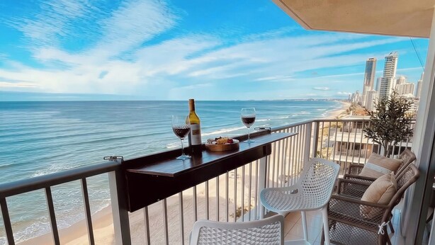 Een balkon met uitzicht op het strand en de oceaan, twee witte stoelen, een tafel met een fles en glazen, en een vloerkleed met een patroon.