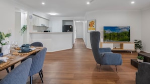 Un salon moderne doté d’un téléviseur à écran plat, d’un coin repas avec une table en bois et d’une cuisine équipée de meubles blancs.