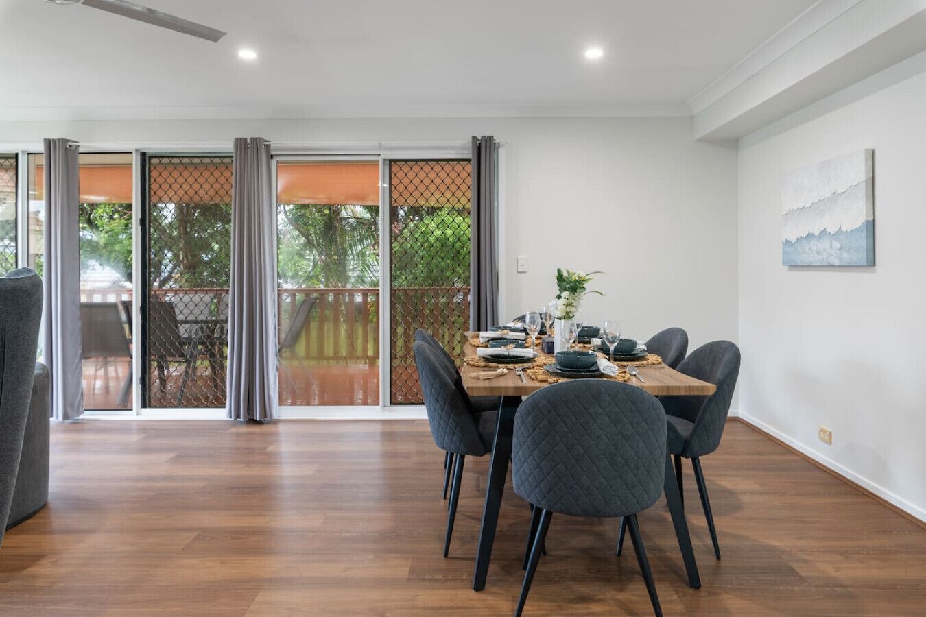 A dining area with a table set for four, chairs, a view through a glass door to an outdoor deck, and a painting on the wall.