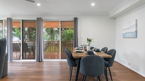 Un coin repas avec une table dressée pour quatre personnes, des chaises, une vue sur une terrasse extérieure à travers une porte vitrée et un tableau accroché au mur.