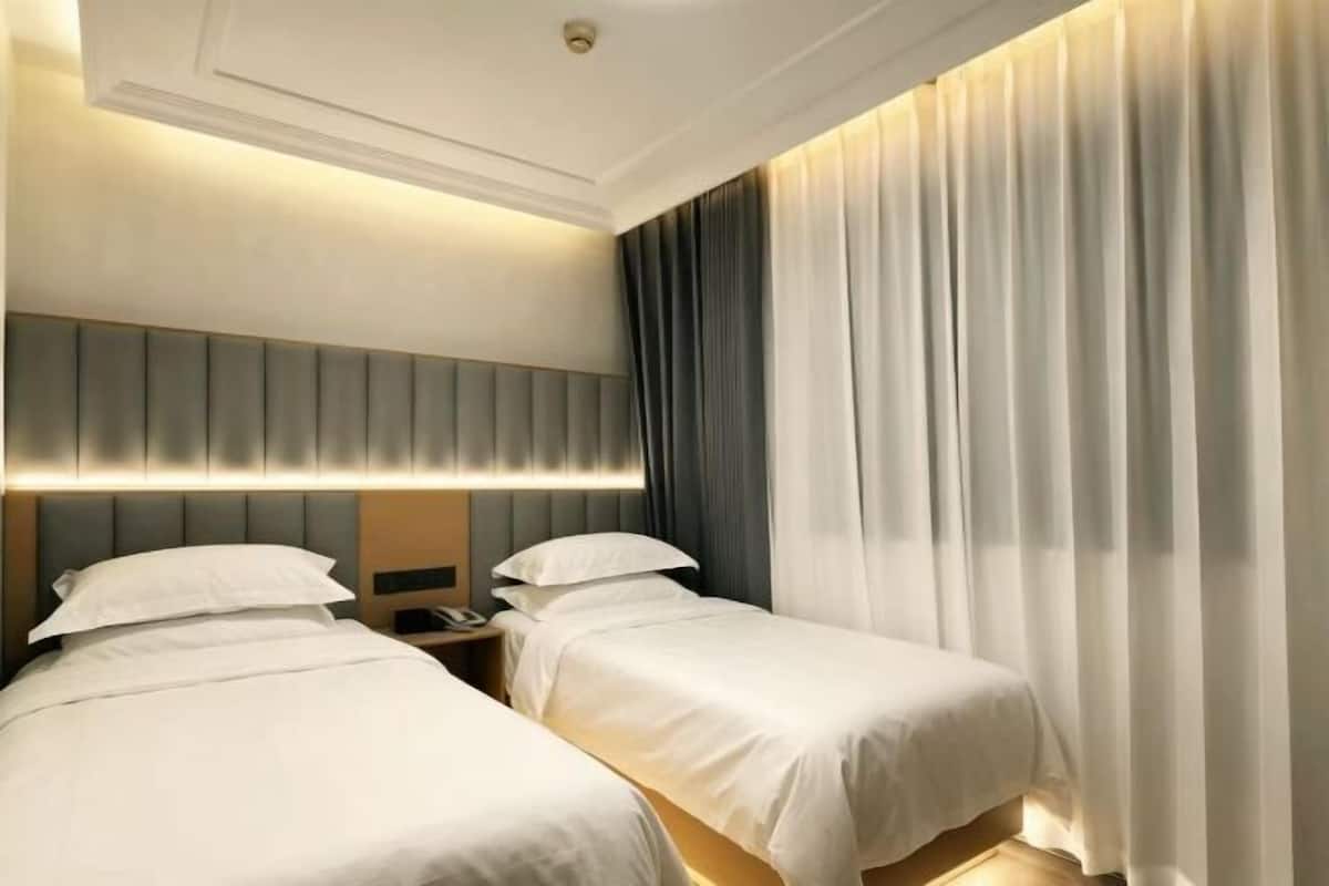 A hotel room with two beds, a headboard with integrated lighting, a bedside table with a phone, and a large window with curtains.