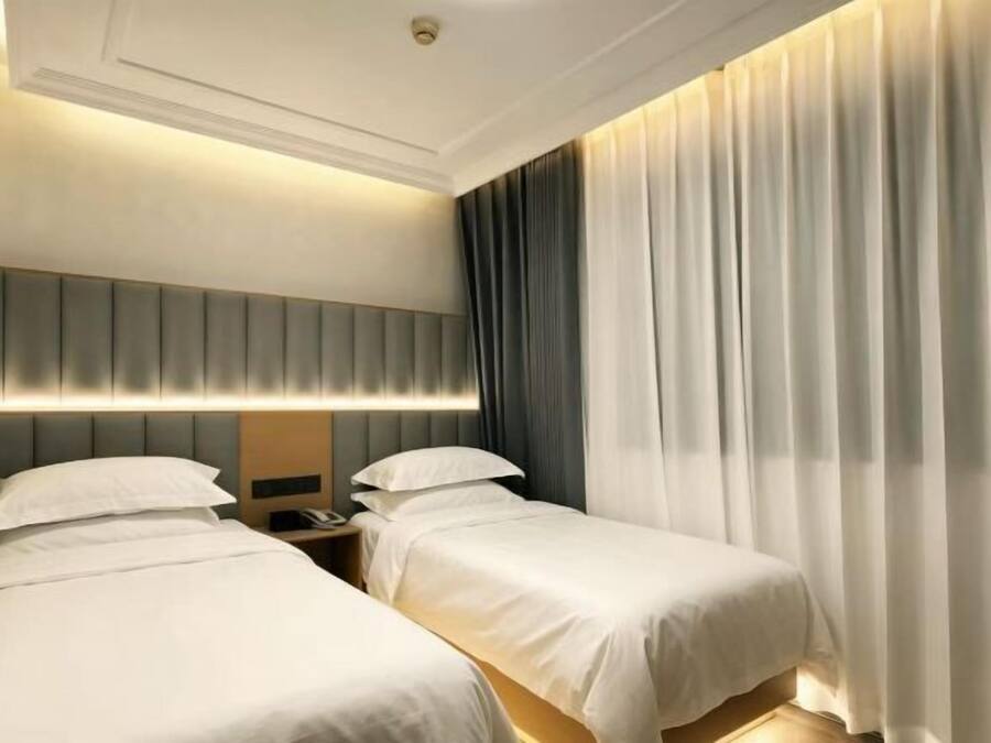 Quarto de hotel com duas camas, cabeceira com iluminação integrada, mesa de cabeceira com telefone e uma janela grande com cortinas.