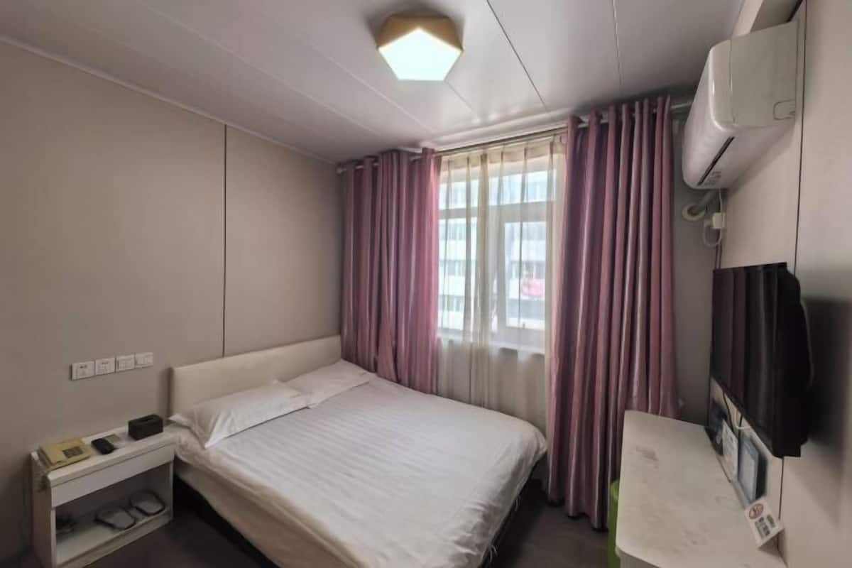 A hotel room with a bed, bedside table, air conditioning unit, and a window with curtains.
