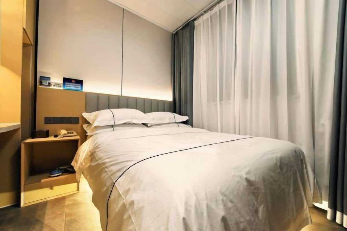 A modern hotel room with a neatly made bed, a bedside table with a phone and a small shelf, and a large window with curtains.