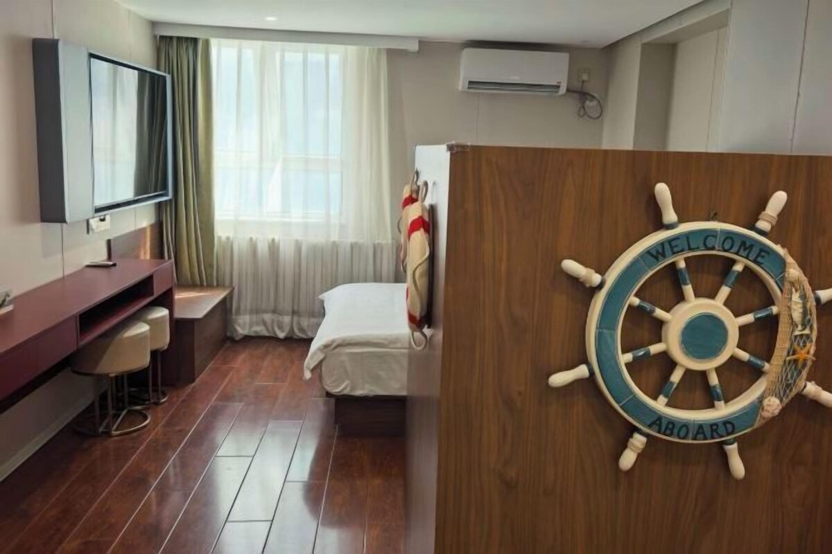 Habitación de hotel con cama, televisor, escritorio con sillas, ventana con cortinas y una decoración con timón de barco.
