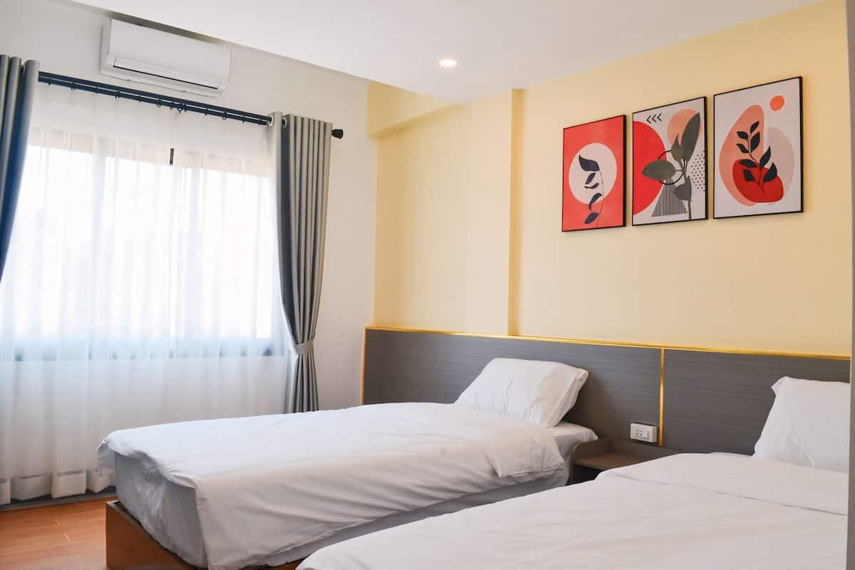A hotel room with two beds, a window with curtains, an air conditioning unit, and three wall-mounted artworks.