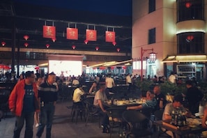 An outdoor dining area with multiple tables and chairs, a stage in the background, and red lanterns hanging above.