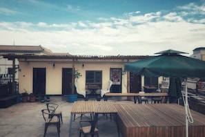 An outdoor patio with wooden tables and chairs, a green umbrella, and a building with a tiled roof in the background. - up north hostel (Dunhuang)
