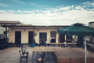 Un patio al aire libre con mesas y sillas de madera, un paraguas verde y un edificio con tejado de baldosas al fondo.