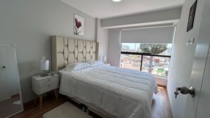 A bedroom with a bed, bedside table, lamp, and a window with a city view.