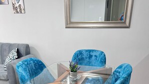 A dining area with a glass-top table, four blue upholstered chairs, a small potted plant, and a grey sofa in the background.