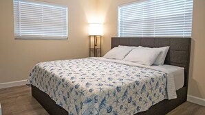 A bedroom with a bed, two windows with blinds, a nightstand with a lamp, and a wooden floor.