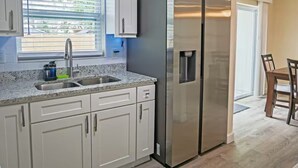 A modern kitchen with white cabinets, a stainless steel refrigerator, a granite countertop, and a window with blinds.