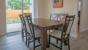 A dining area with a wooden table and chairs, a sliding glass door leading to a backyard, and a sunset painting on the wall.
