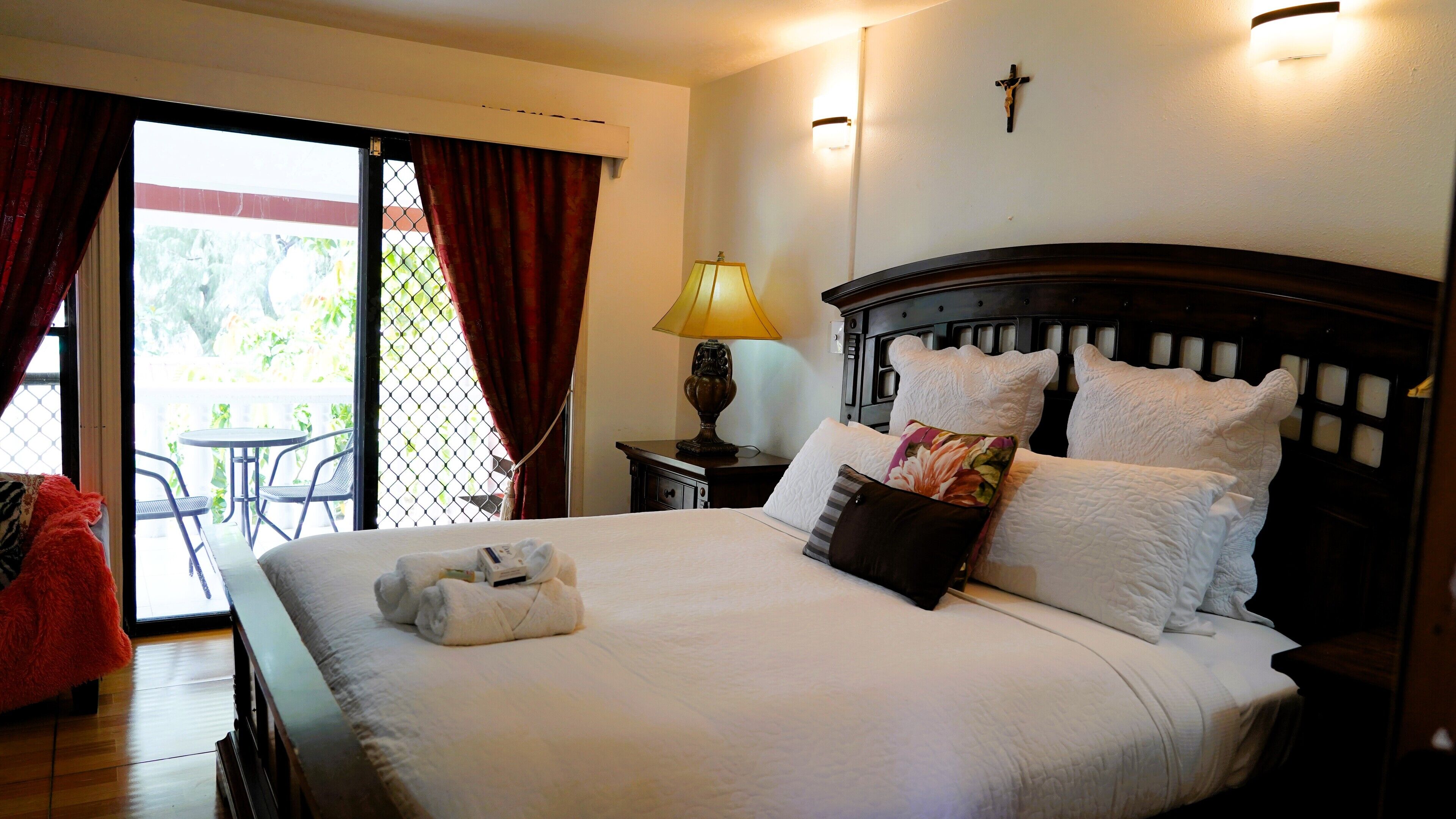 Een netjes opgemaakt bed met witte lakens en een bloemenkussen, een bedlampje en een kruis aan de muur.