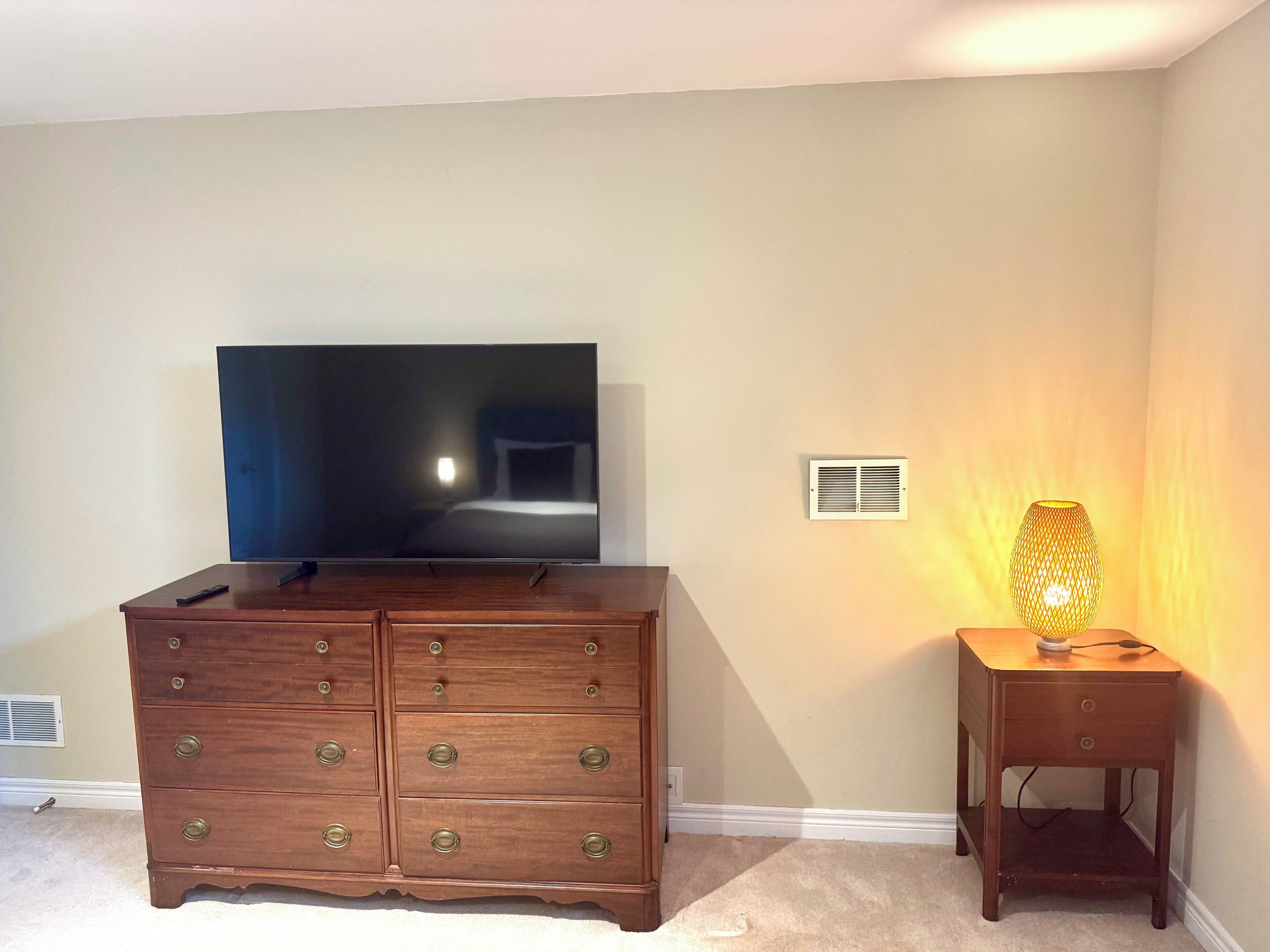 A room with a large flat-screen TV on a wooden dresser, a small table with a lamp, and a vent on the wall.