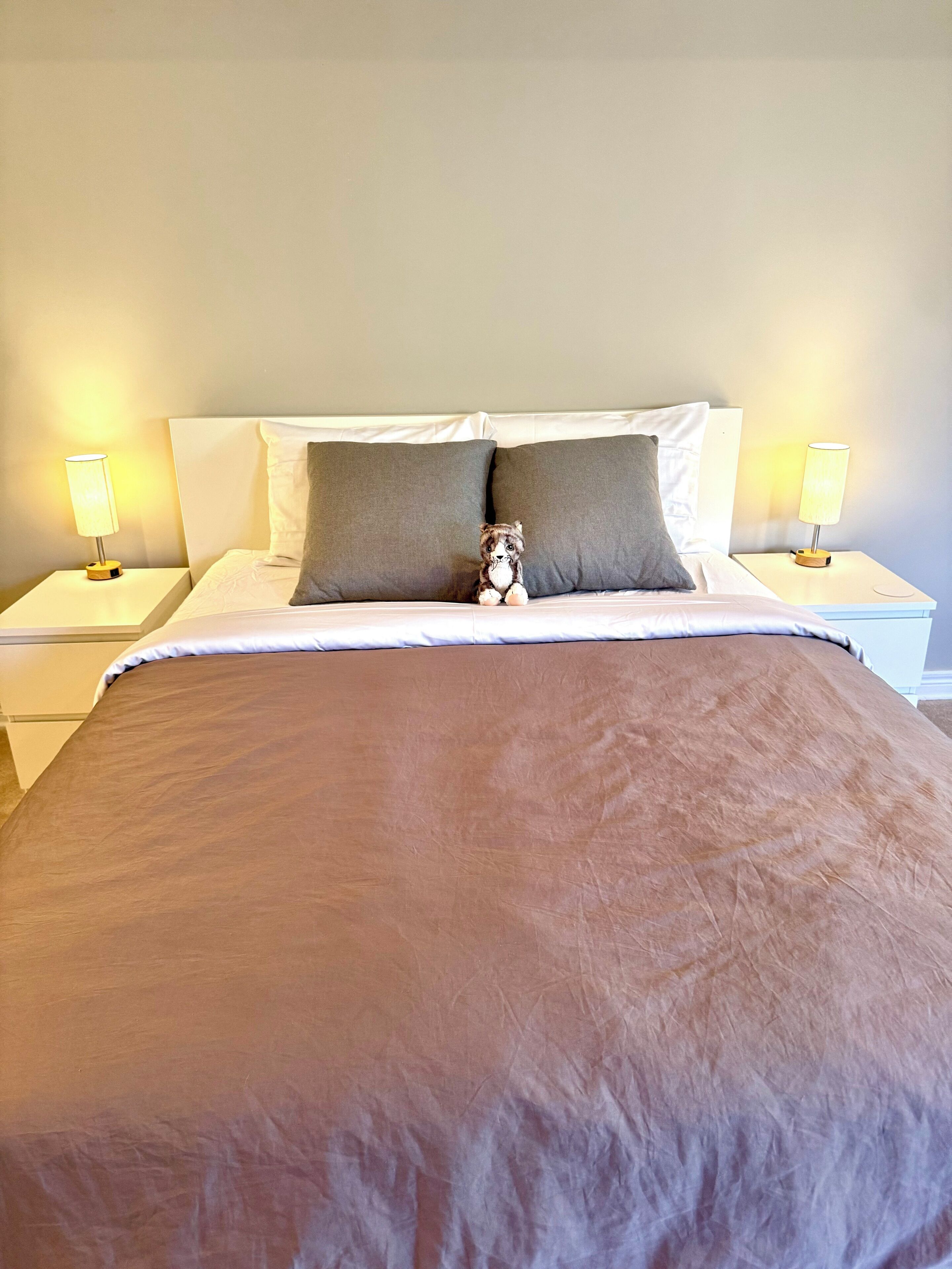 Una cama bien hecha con una colcha marrón, dos almohadas y un pequeño peluche. Hay dos lámparas de noche en mesitas blancas.
