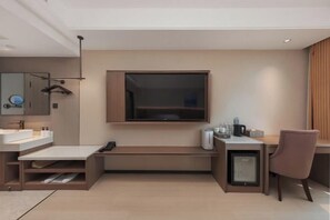 A modern hotel room with a large flat-screen TV mounted on the wall, a small desk with a chair, a minibar, and a large mirror.