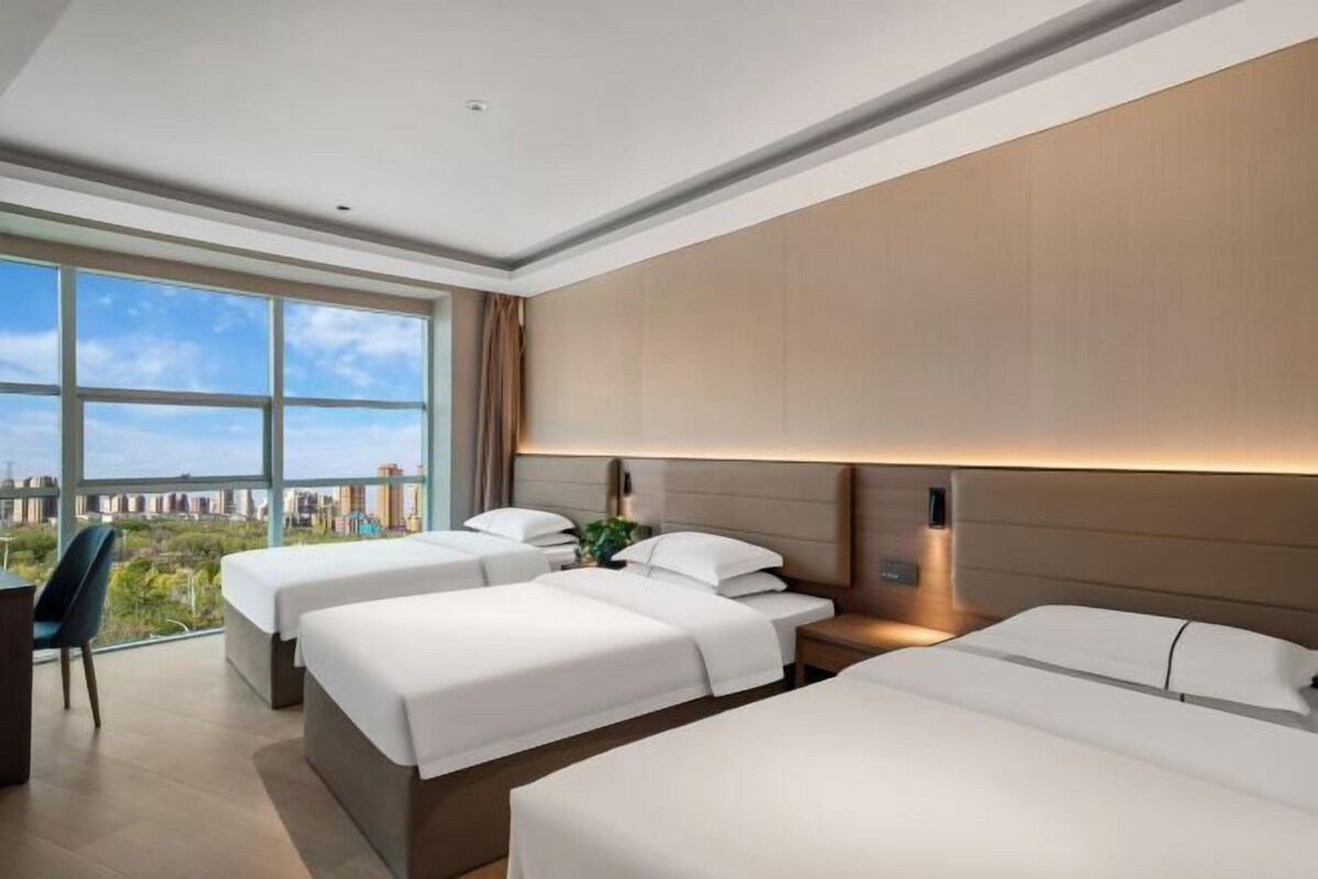 Una habitación de hotel moderna con tres camas, un ventanal amplio con vista a la ciudad y un escritorio con una silla.