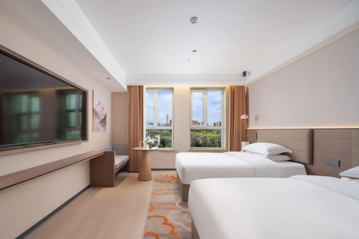 Habitación de hotel con dos camas, un ventanal amplio, una mesita pequeña y vistas a la ciudad.
