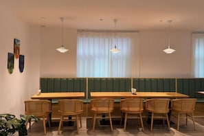 A dining area with wooden tables and chairs, pendant lighting, and a view through a window with sheer curtains.