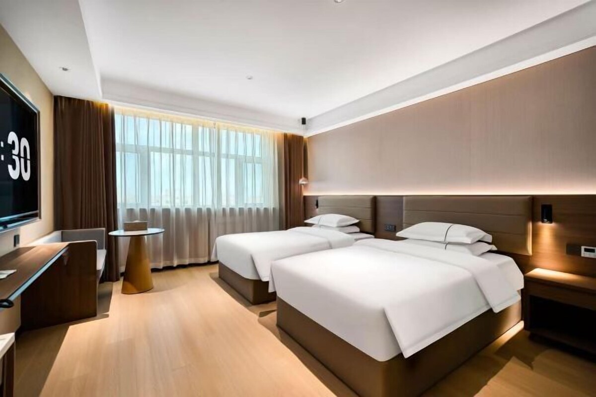 Una habitación de hotel moderna con dos camas, televisión, una mesita y un gran ventanal con cortinas.